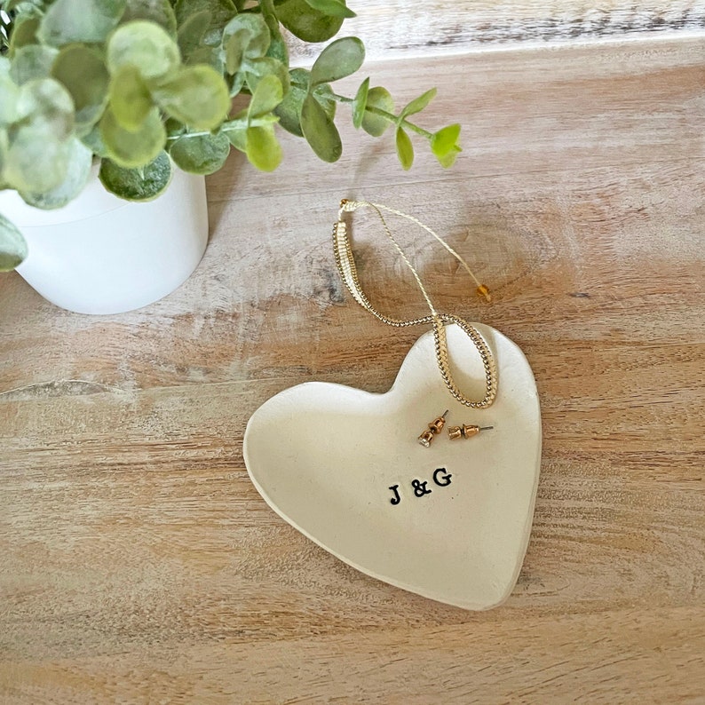 May include: A heart-shaped, off-white ceramic jewelry dish with the initials "J & G" stamped in black. A gold necklace and a pair of stud earrings rest on the dish. A small white pot with greenery is in the background.