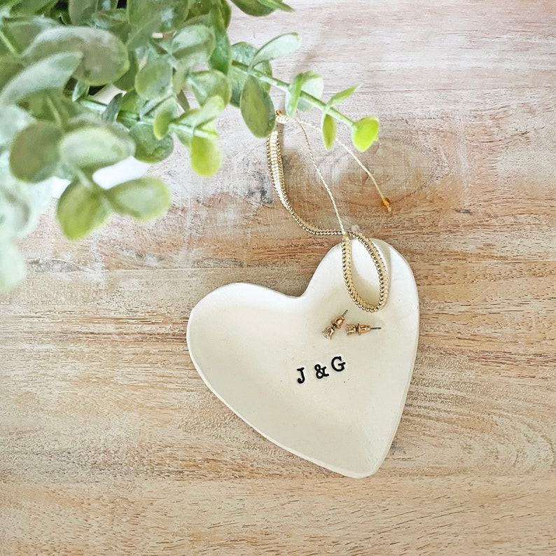 May include: A heart-shaped, off-white ceramic trinket dish with the initials "J & G" stamped in black. A gold necklace and small stud earrings rest on the dish. Greenery is visible in the background.