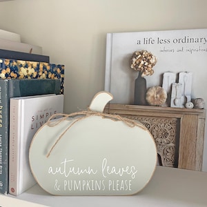 May include: A light sage green pumpkin-shaped wooden sign with a natural jute twine bow tied around the stem. The text "autumn leaves & PUMPKINS PLEASE" is printed in white script and block lettering on the front of the smooth, flat surface.