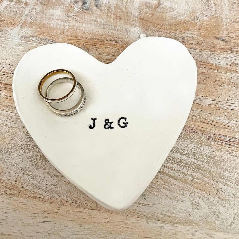 May include: A white heart-shaped ring dish with the initials "J & G" stamped on it. Two rings, one gold and one silver with small stones, rest on the dish. The dish sits on a light wood surface.