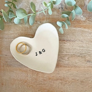 May include: Heart-shaped, off-white ceramic ring dish with the initials "J & G" stamped in black. Two rings, one gold and one silver, rest inside the dish. The dish sits on a light-colored wooden surface, with green foliage in the background.