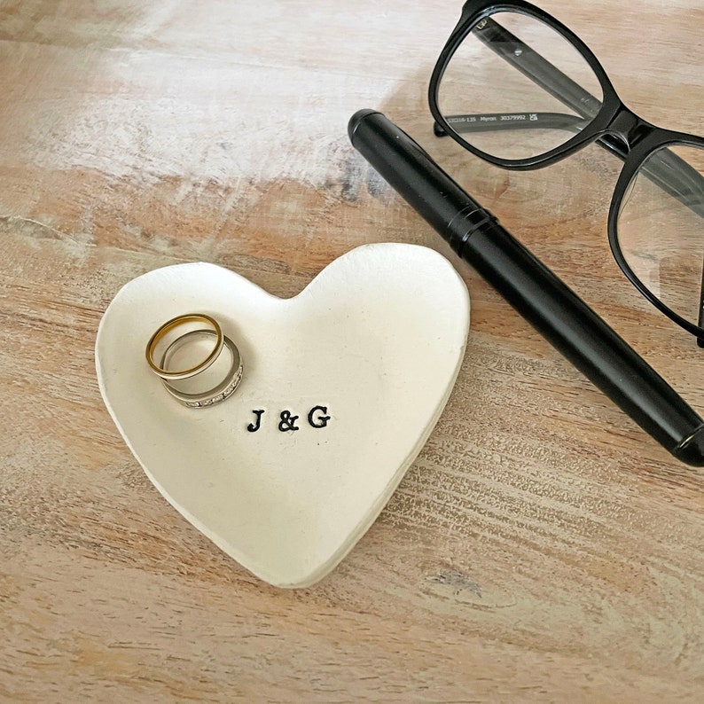 May include: A white heart-shaped trinket dish with the initials "J & G" stamped in black. Two rings rest inside the dish. A black pen and black-framed glasses are also on the wooden surface.