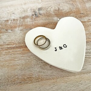 May include: A white heart-shaped ceramic ring dish with the initials "J & G" in black. Two rings, one gold and one silver with clear stones, rest on the dish. The dish sits on a light-colored wooden surface.