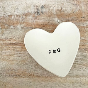 May include: A white heart-shaped trinket dish with the stamped letters "J & G" in black. The dish is set on a light brown wooden surface, creating a rustic and romantic aesthetic.