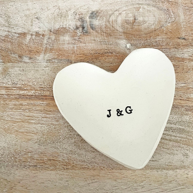 May include: A white heart-shaped trinket dish with the stamped letters "J & G" in black. The dish is set on a light brown wooden surface, creating a rustic and romantic aesthetic.
