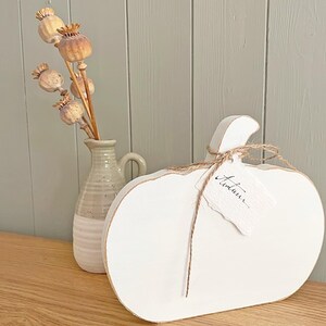 May include: A flat, white wooden pumpkin cutout with a jute twine bow and a small, rectangular paper tag featuring the handwritten word "Autumn" in black ink. Beside it stands a two-toned ceramic pitcher holding four dried poppy seed pods.