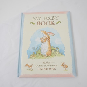Puede incluir: Un libro para bebés titulado "MY BABY BOOK" con un borde azul claro y rosa. La portada presenta una ilustración en acuarela de un conejo marrón sosteniendo un conejo más pequeño. El texto "Based on Guess How Much I Love You" también es visible.