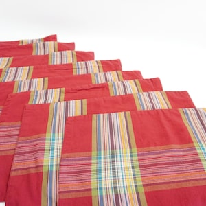 May include: A set of red placemats with a plaid pattern. Each placemat features a vibrant red background with colorful stripes in yellow, blue, green, and purple. The placemats are arranged in a cascading display, showcasing their design.
