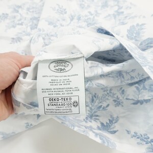 May include: White fabric with a blue floral pattern. The fabric is held up to reveal a tag with text including "MADE IN INDIA" and "OEKO-TEX STANDARD 100". The fabric is likely a sheet or other bedding item.