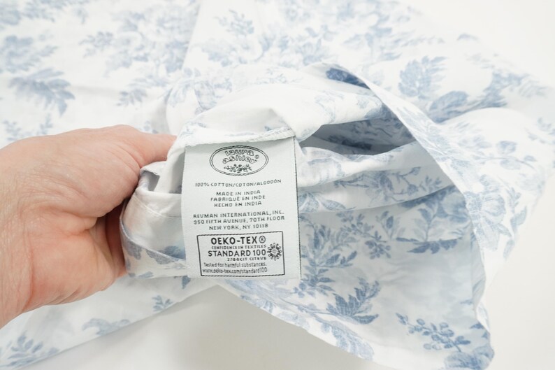 May include: White fabric with a blue floral pattern. The fabric is held up to reveal a tag with text including "MADE IN INDIA" and "OEKO-TEX STANDARD 100". The fabric is likely a sheet or other bedding item.