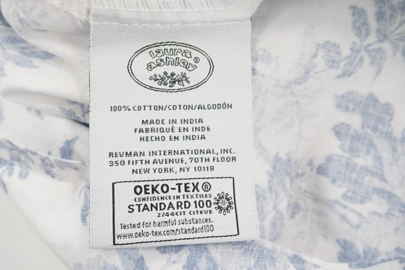 May include: A white fabric tag with the "Laura Ashley" logo, text indicating 100% cotton, and "Made in India." The tag also includes the OEKO-TEX Standard 100 certification and the address of Revman International in New York.
