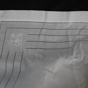 May include: White linen textile with embroidered floral and linear designs. The textile features a delicate floral motif in the corner and parallel lines across the surface. The edges are neatly finished, suggesting a decorative or functional item.