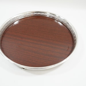 May include: A circular serving tray with a silver-coloured rim and a dark brown wooden centre. The rim has a decorative pattern. The tray is on a white background. Suitable for serving drinks or snacks.