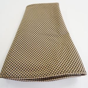 May include: Folded fabric with a tan and black checkered pattern. The fabric is folded in a triangular shape, with the checkered pattern visible on the top surface. The edges of the fabric are neatly finished.