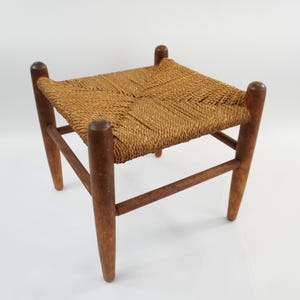 May include: A small wooden stool featuring four tapered legs and a square seat woven from natural tan fibre cord. The frame is constructed from medium-toned wood with a smooth finish, and the woven seat displays a distinct diagonal pattern.