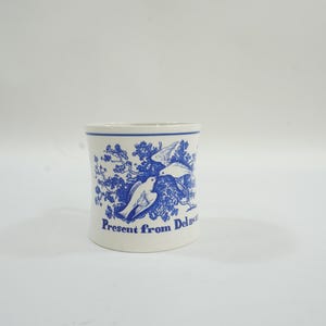 May include: A white ceramic mug with a blue floral and bird design. The mug has a cylindrical shape and features the text "Present from Delaware" in blue. The design includes two birds and branches.