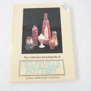 Puede incluir: Un libro titulado "The Collectors Encyclopedia of Roseville Pottery" con una cubierta crema. La portada presenta cerámica Roseville en rojo, rosa y blanco. El libro es una segunda serie de Sharon y Bob Huxford. Se ve la palabra "Values Updated".