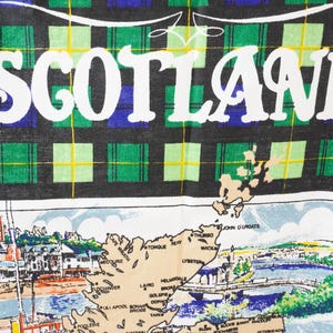 May include: A textile featuring the word "SCOTLAND" in white, set against a black, green, blue, and yellow plaid pattern. Below, a map of Scotland is depicted with illustrations of landmarks and cities.