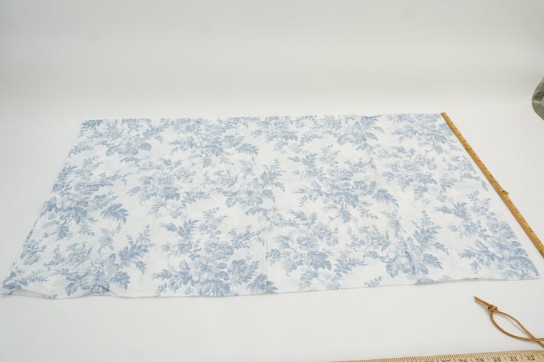 May include: A rectangular piece of fabric with a white background and a blue floral pattern. A wooden ruler is placed along the edge of the fabric. The fabric is likely used for sewing or crafting.