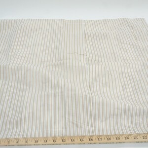 May include: Cream-colored fabric with vertical stripes in a light brown hue. A wooden ruler with inch markings is placed along the bottom edge. The fabric is likely used for sewing or crafting projects.