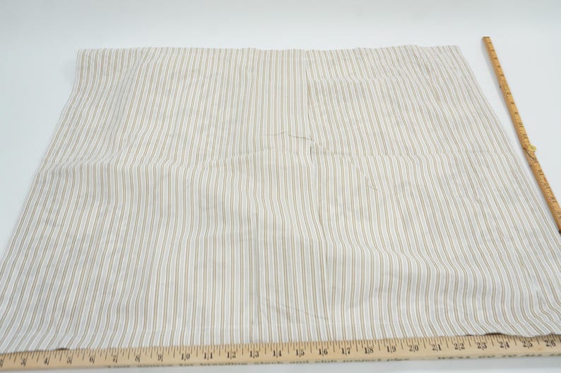 May include: Cream-colored fabric with vertical stripes in a light brown hue. A wooden ruler with inch markings is placed along the bottom edge. The fabric is likely used for sewing or crafting projects.