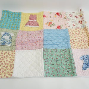 Puede incluir: Un edredón de retazos con varias telas florales y estampadas en colores pastel. Vestidos aplicados están cosidos en el edredón. El edredón mide aproximadamente 127 cm de ancho y 152 cm de largo.