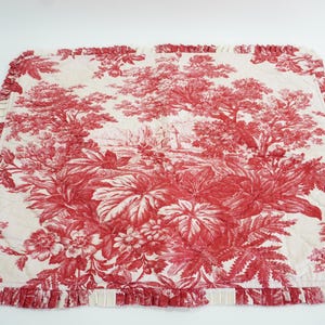 May include: A white fabric pillow cover with a red floral pattern. The pattern features a toile design with trees, flowers, and leaves. The pillow cover has a ruffled edge.