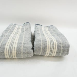 May include: Two folded gray and white striped quilted fabric items. The fabric has a light gray background with vertical white stripes. The edges are finished with a dark blue trim. The items are placed side by side on a white surface.