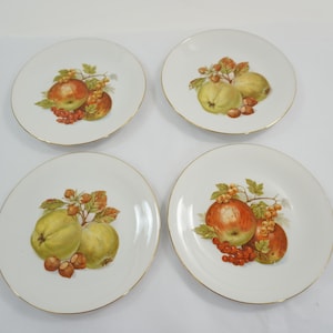 以下が含まれることがあります： 金色の縁取りが施された4つの白いセラミックプレート。それぞれに果物の静物画が描かれています。果物にはリンゴ、梨、赤いベリーが含まれており、緑と茶色の葉が付いています。プレートは正方形に配置されています。