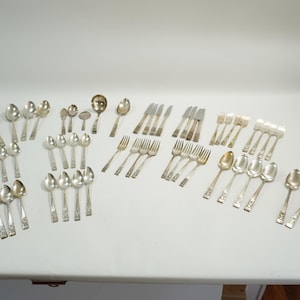May include: A large collection of silver-toned metal flatware including various sizes of spoons, forks, and knives. The set features multiple groupings of matching utensils with decorative handles, arranged in rows on a plain white surface.