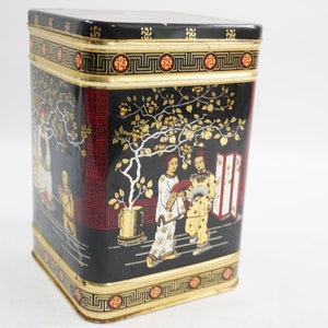 Puede incluir: Lata de té de metal negro y dorado con un diseño de inspiración china que presenta un árbol, dos figuras y una pantalla roja y blanca. La lata tiene un borde dorado y un borde dorado.