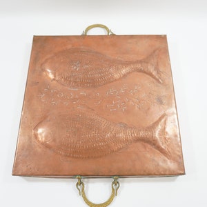 Peut inclure: Plateau de service rectangulaire en cuivre avec deux poissons en relief et des motifs décoratifs. Il possède des poignées en laiton et une couleur brun-rouge. Une pièce unique pour servir ou décorer, environ 30 cm de côté.