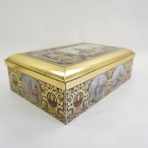 May include: Rectangular gold-colored tin box with a hinged lid. The box features ornate detailing with images of buildings and landscapes. The sides and lid are decorated with intricate patterns and circular vignettes.