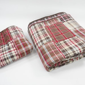 Könnte beinhalten: Zwei gefaltete Steppdecken mit einem Patchwork-Design aus kariertem Stoff in den Farben Rot, Braun, Weiß und Hellblau. Die Decken haben einen dunkelbraunen Rand und sichtbare Nähte, die eine gesteppte Textur auf der Stoffoberfläche erzeugen.