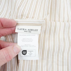 May include: Close-up of a fabric with vertical stripes in beige and white. A label with the text "LAURA ASHLEY SINCE 1953" is visible, along with other text and the OEKO-TEX STANDARD 100 logo. The fabric is held by a hand.