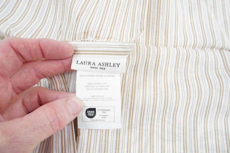 May include: Close-up of a fabric with vertical stripes in beige and white. A label with the text "LAURA ASHLEY SINCE 1953" is visible, along with other text and the OEKO-TEX STANDARD 100 logo. The fabric is held by a hand.