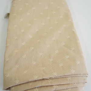 May include: Folded beige fabric featuring a delicate vertical stripe and small, repeating leaf motifs. The fabric is neatly stacked, highlighting its texture and colour. The neutral tone suggests a versatile material suitable for various textile projects.