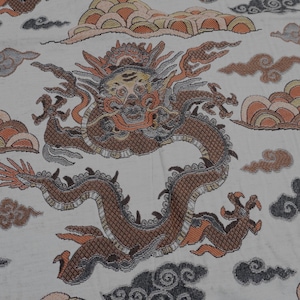 May include: A white fabric featuring a repeating pattern of a brown dragon with intricate scales and a detailed face. The dragon is surrounded by stylized clouds in shades of orange, yellow, and dark gray, creating a textured, woven appearance.
