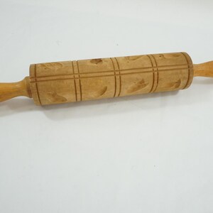 May include: A wooden rolling pin with a decorative pattern. The rolling pin is light brown with a series of carved designs and wooden handles. The rolling pin measures approximately 12 inches in length.