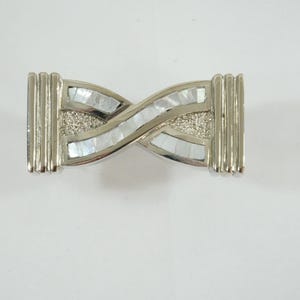 Könnte beinhalten: Silberfarbene Metallschnalle mit einem verschlungenen Design. Die Schnalle hat Bereiche aus schillerndem Perlmutt und einen zentralen Bereich mit strukturiertem, glitzerndem Aussehen. Die Enden haben vertikale Rippendetails.