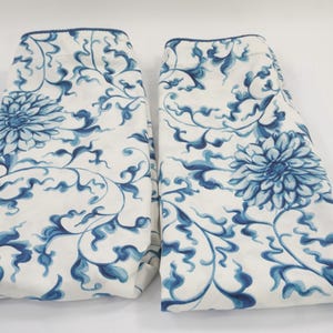 Peut inclure: Deux panneaux de tissu rectangulaires présentant une base blanche avec un motif répétitif de fleurs bleues et de vignes entrelacées. Chaque panneau possède un bord supérieur fini avec une fine bordure bleue, affichant un motif botanique.