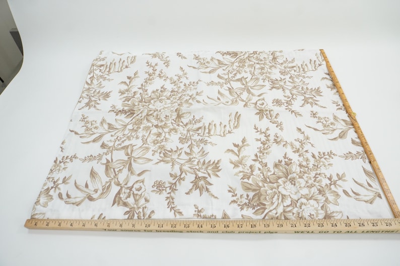 May include: A piece of fabric with a floral pattern in shades of brown and beige. The design features detailed botanical illustrations of flowers and foliage. A wooden ruler with inch measurements is placed along the edge of the fabric.