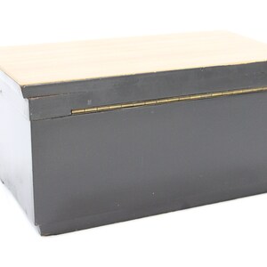 May include: A black wooden box with a hinged lid and a brass hinge. The lid is made of a light wood.