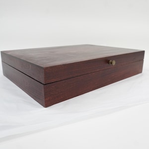 Puede incluir: Una caja de almacenamiento rectangular de madera de color marrón oscuro con un acabado liso y un pequeño pomo circular de latón centrado en el borde frontal. La caja presenta un diseño de tapa con bisagras, bordes rectos y superficie plana.