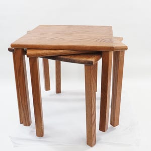 May include: Set of three nesting tables made of light brown wood. The tables have square tops with rounded corners and straight legs. The tables are stacked together, with the smallest table nested underneath the larger ones.