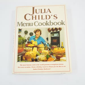 以下が含まれることがあります： Julia Child's Menu Cookbookというタイトルのハードカバー本。白い表紙には、青いシャツを着た人物が様々な料理、調理器具、吊るされた鍋に囲まれている写真が印刷されており、下部には説明文が記載されています。