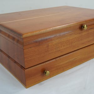 Puede incluir: Una caja de madera con un acabado marrón rojizo cálido. La caja tiene una tapa con bisagras y un cajón, ambos con pomos de color latón. La veta de la madera es visible, lo que se suma a la estética natural de la caja. La caja probablemente se utiliza para el almacenamiento.