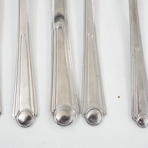 May include: Close-up of five silver-colored forks. The forks have a rounded base with a circular design and a tapered handle with vertical lines. The forks are arranged on a white surface.