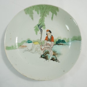 以下が含まれることがあります： 木の下にいる人物の手描きのシーンが描かれた白いセラミックプレート。人物は赤いトップスを着ており、水辺の近くに座っています。緑と青のアクセントが風景デザインに使用されています。