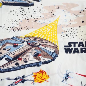 Op de afbeelding: Wit Star Wars-thema stof met de Millennium Falcon, X-wing fighters en het Star Wars-logo. Het ontwerp bevat een gele uitbarsting en een ruimteachtergrond met zwarte stippen.